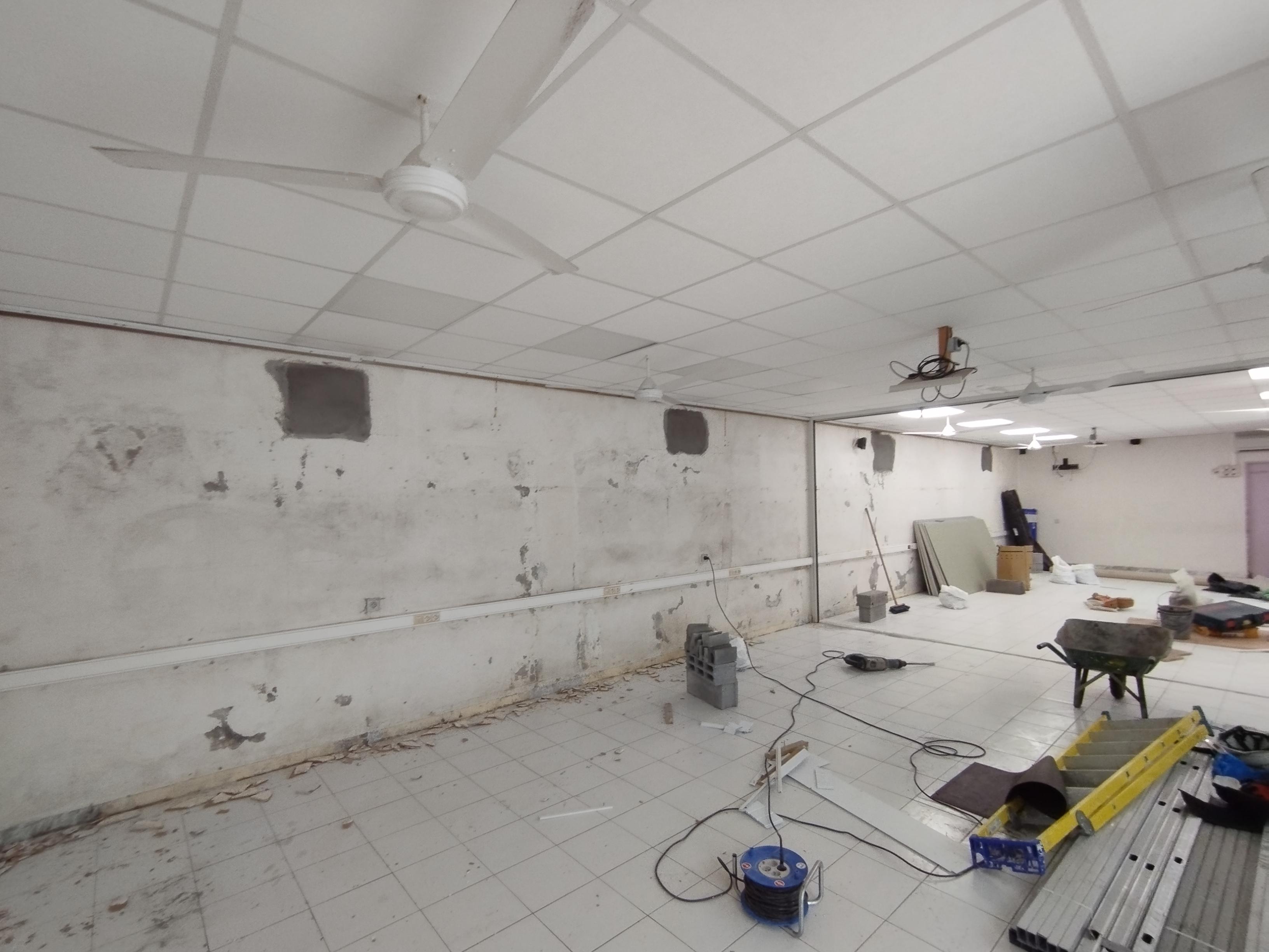 Pièce spacieuse et inachevée avec un sol carrelé blanc et des murs endommagés. Des ventilateurs de plafond sont suspendus à un plafond à grille. Des outils et des matériaux de construction sont éparpillés.