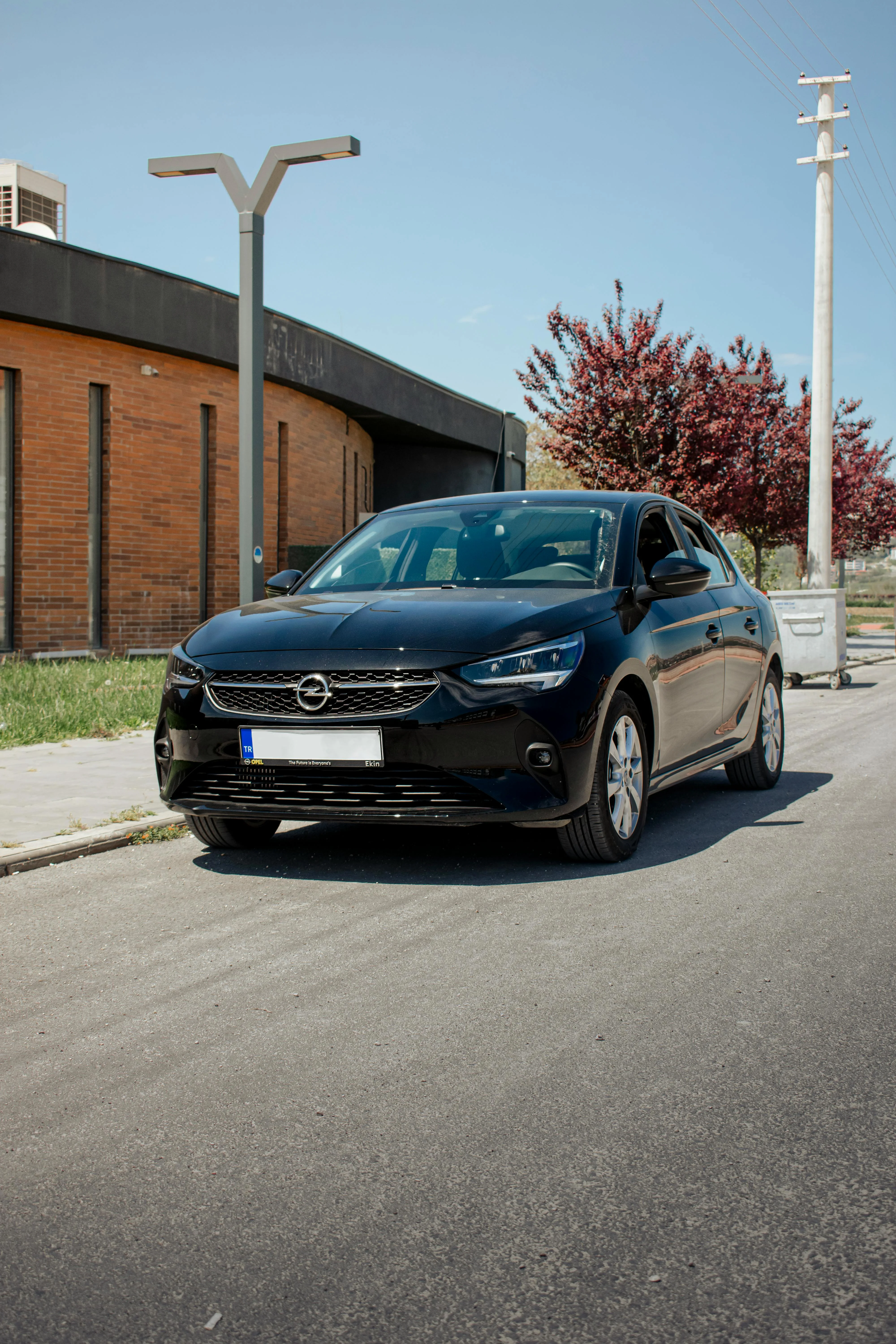 Opel Corsa noire photographiée sur une route urbaine sous un ciel clair, mettant en avant la nouvelle identité visuelle de la marque avec sa calandre élégante, ses lignes tendues et son style sobre mais moderne. Une scène lumineuse qui valorise la silhouette compacte et contemporaine de la citadine Opel.