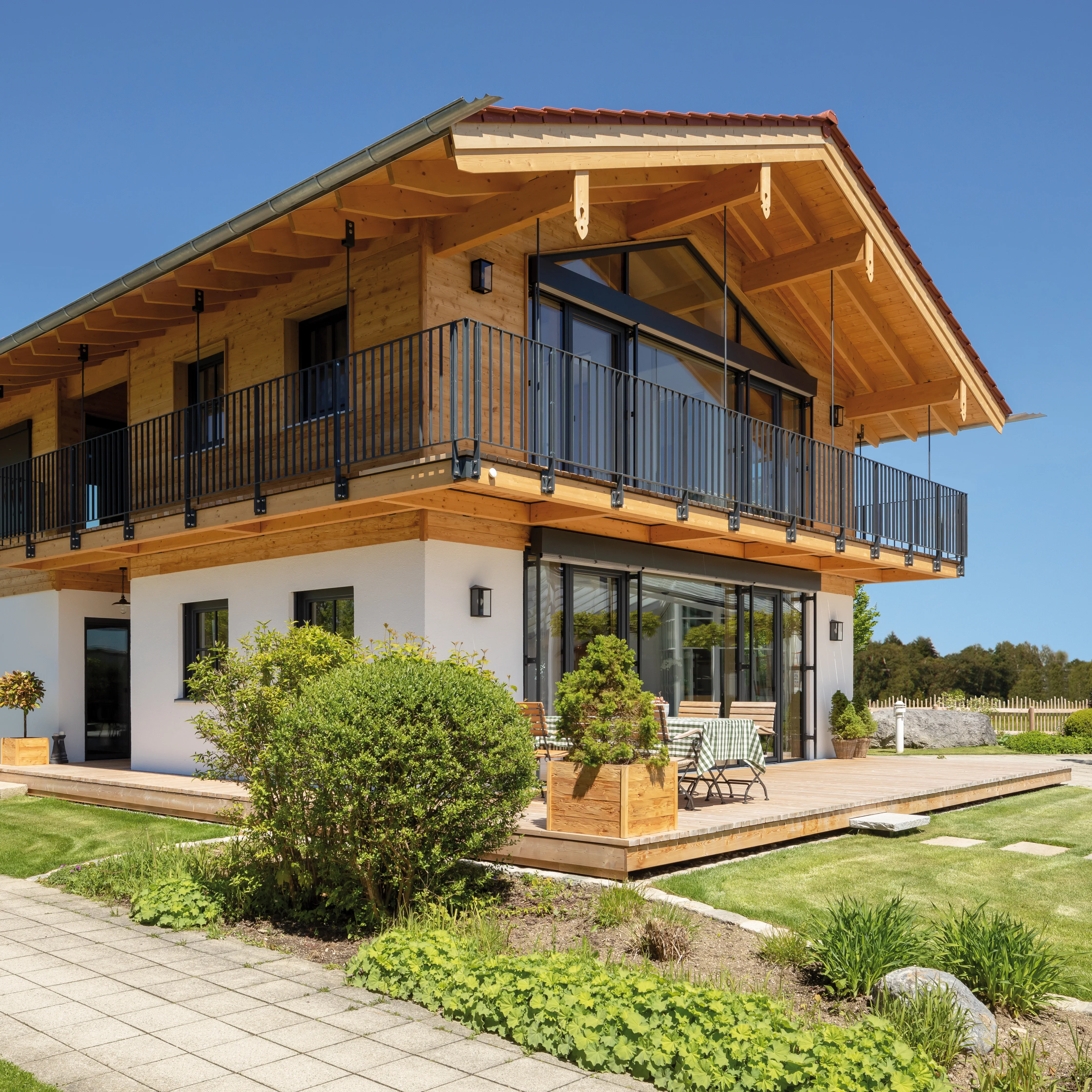 Modernes Holzhaus mit weißen Wänden, großzügiger Holzterrasse, Balkon und Gartensitzgruppe unter blauem Himmel.