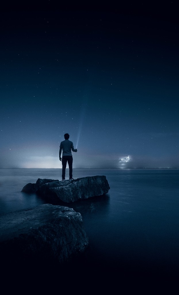 A man shining a light into the sky standing on rocks at the edge of a lake