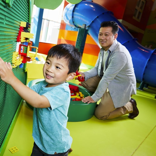 Nasmijano dijete igra se građevnim kockama na zelenom zidu, dok muškarac kleči u blizini i slaže kocke. U pozadini je plavi tobogan.
