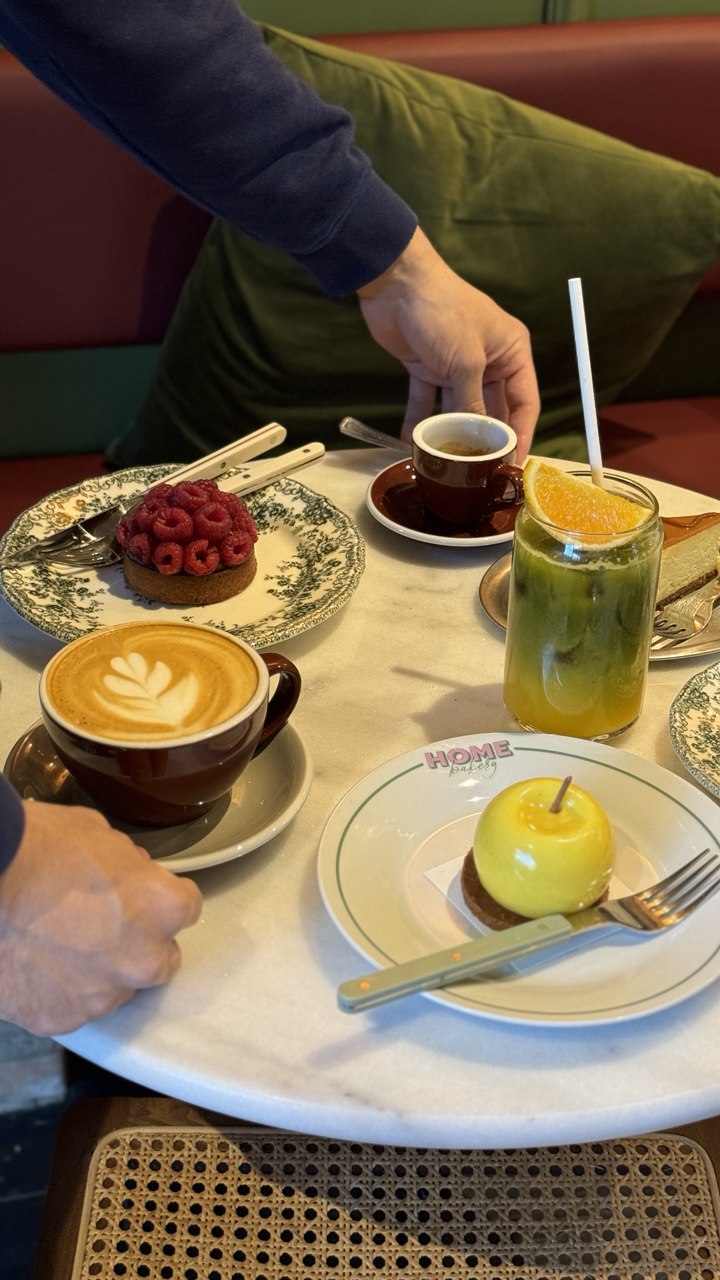A cozy café setting features a marble table with an artistically crafted latte, a small espresso, a vibrant green iced drink with an orange slice, a raspberry-topped pastry, and a decorative sweet resembling a shiny yellow apple.