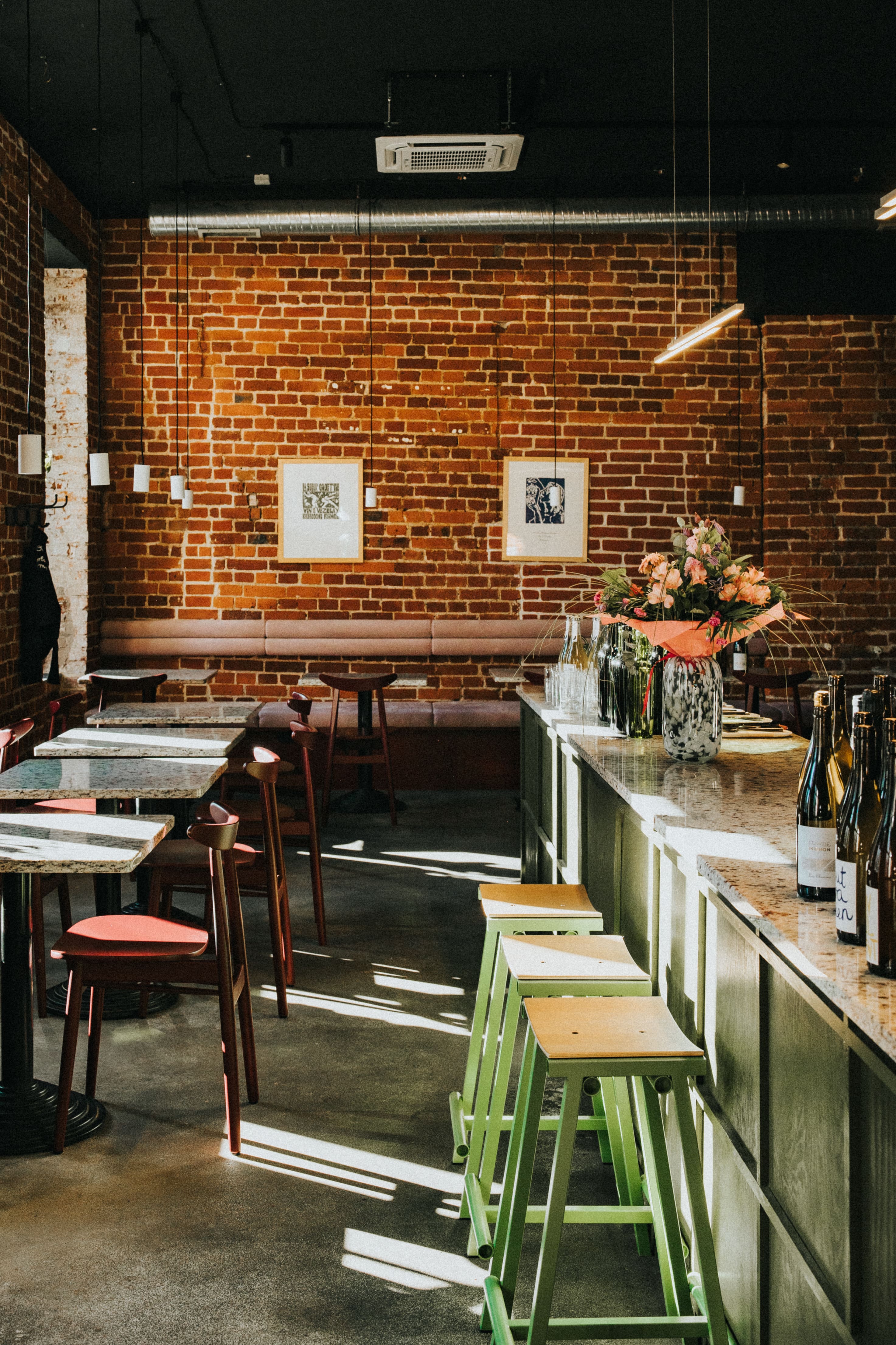 Wnętrze bistro Pijalni z ceglanymi ścianami, oprawioną sztuką i oświetloną słońcem przestrzenią. Zielone stołki przy barze z butelkowanymi napojami, kwiaty dodające ciepłego akcentu.