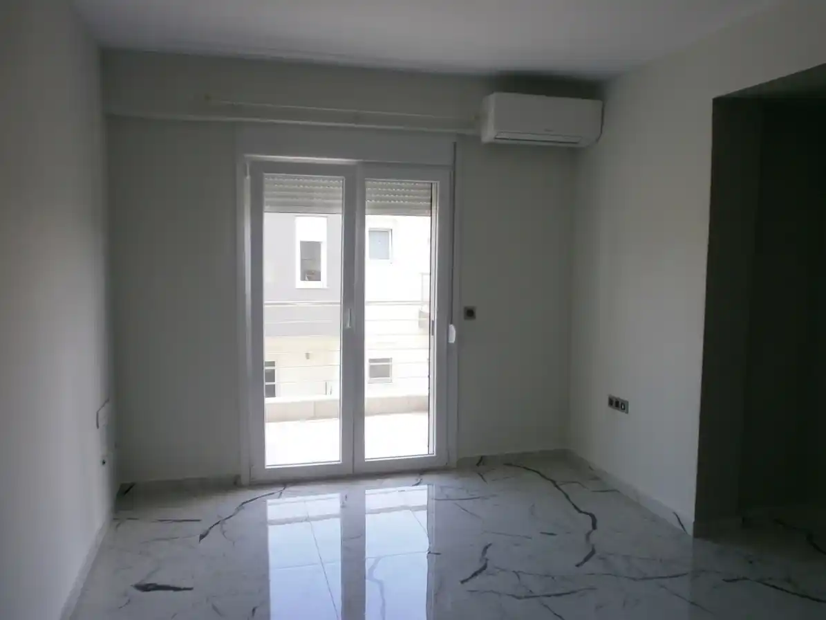 Άδειο σαλόνι με κλιματιστικόμε λευκούς τοίχους, μπαλκονόπορτα και γυαλιστερά πλακάκια σε όψη λευκού μαρμάρου.