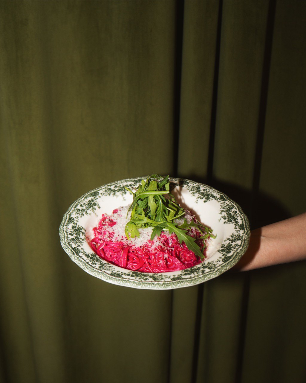 A decorative plate with an intricate border features vibrant red beet ravioli topped with a garnish of fresh greens against a backdrop of dark green curtains.