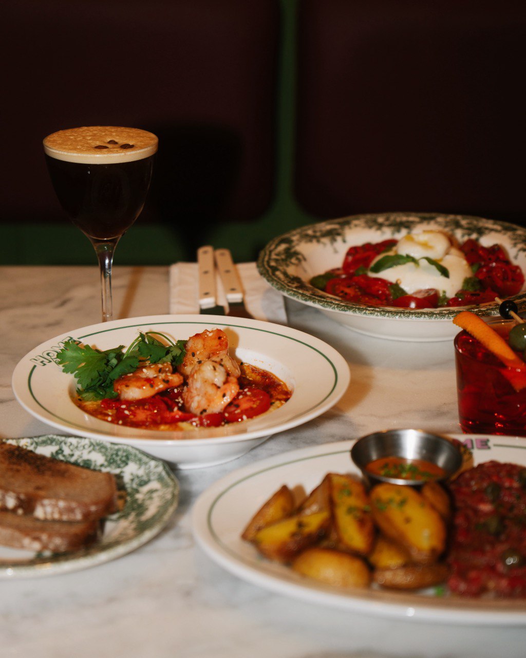 A gourmet dining spread features a dish of marinated shrimp garnished with cilantro, a bowl of fresh burrata and tomatoes, a plate of roasted potato wedges, and a creamy espresso dessert drink on a marble table.