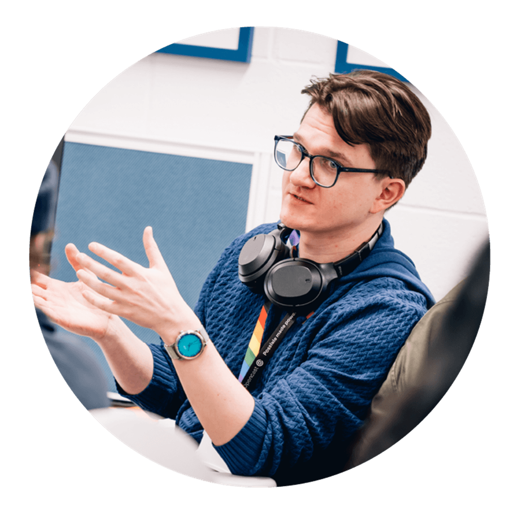 A person with glasses and headphones, gesturing with their hands as he speaks. They wear a blue sweater and a watch.