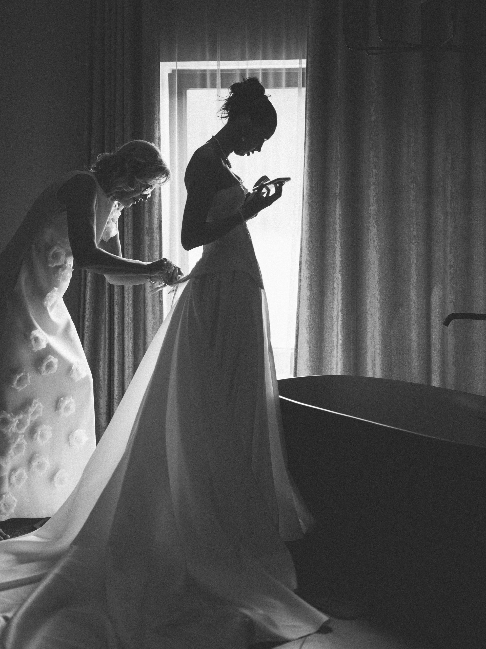 Black-and-white preparation scene with the bride silhouetted at a window while another person adjusts her train.