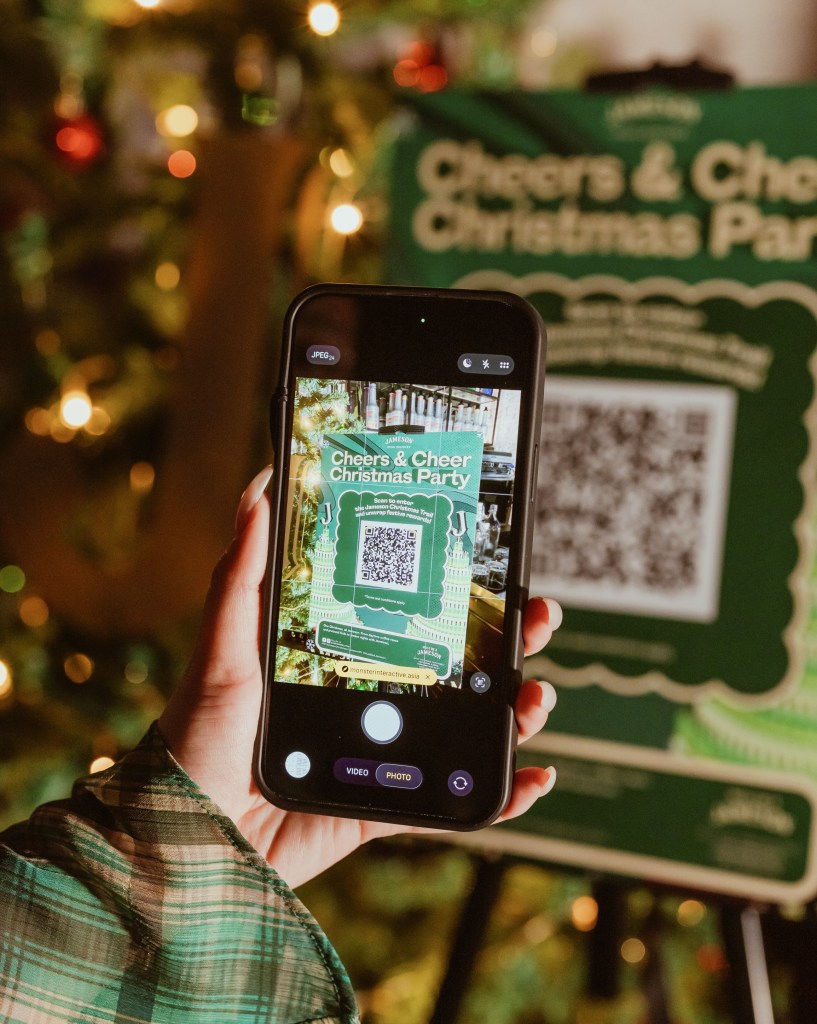 A hand holding a smartphone displaying a QR code for the 'Cheers & Cheer Christmas Party' in front of a festive backdrop with lights.