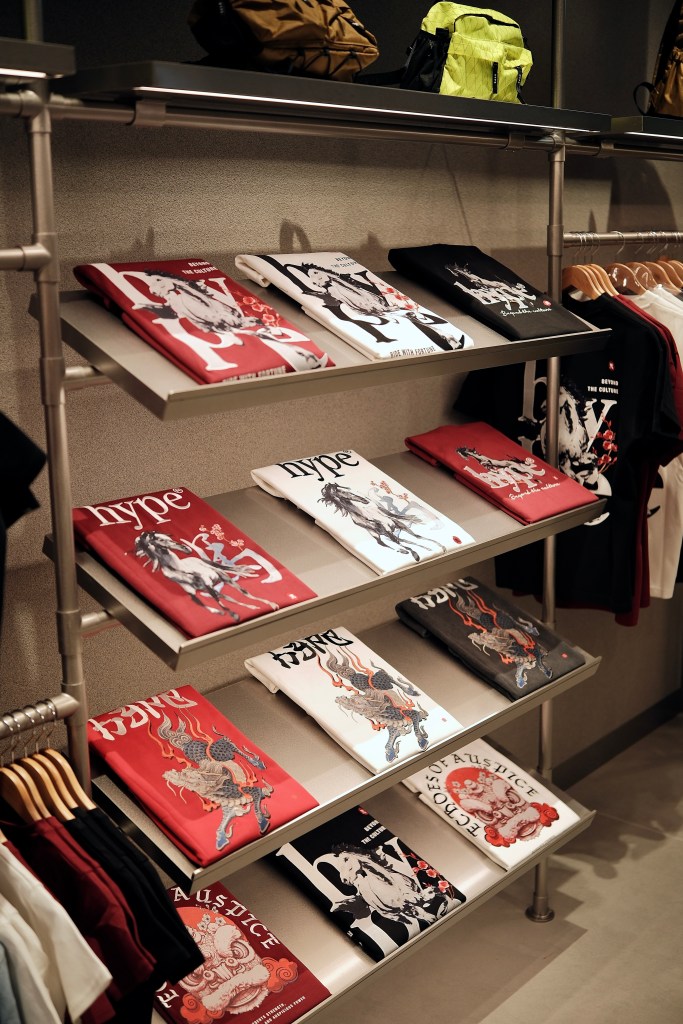 A display of neatly folded t-shirts with various graphic designs in a retail store, featuring red, black, and white colors, alongside backpacks on the top shelf.