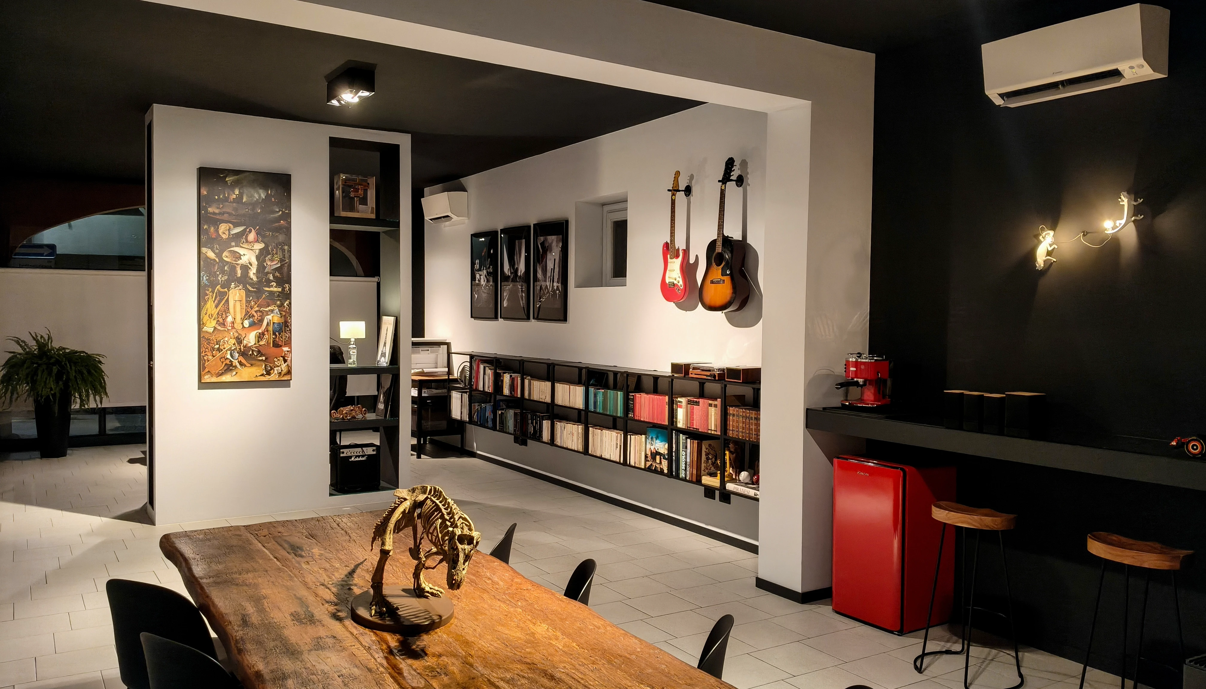 Modern architectural workspace with large floor-to-ceiling windows, surrounded by trees, and desks filled with design materials and books. Two blurred figures are walking through the space.