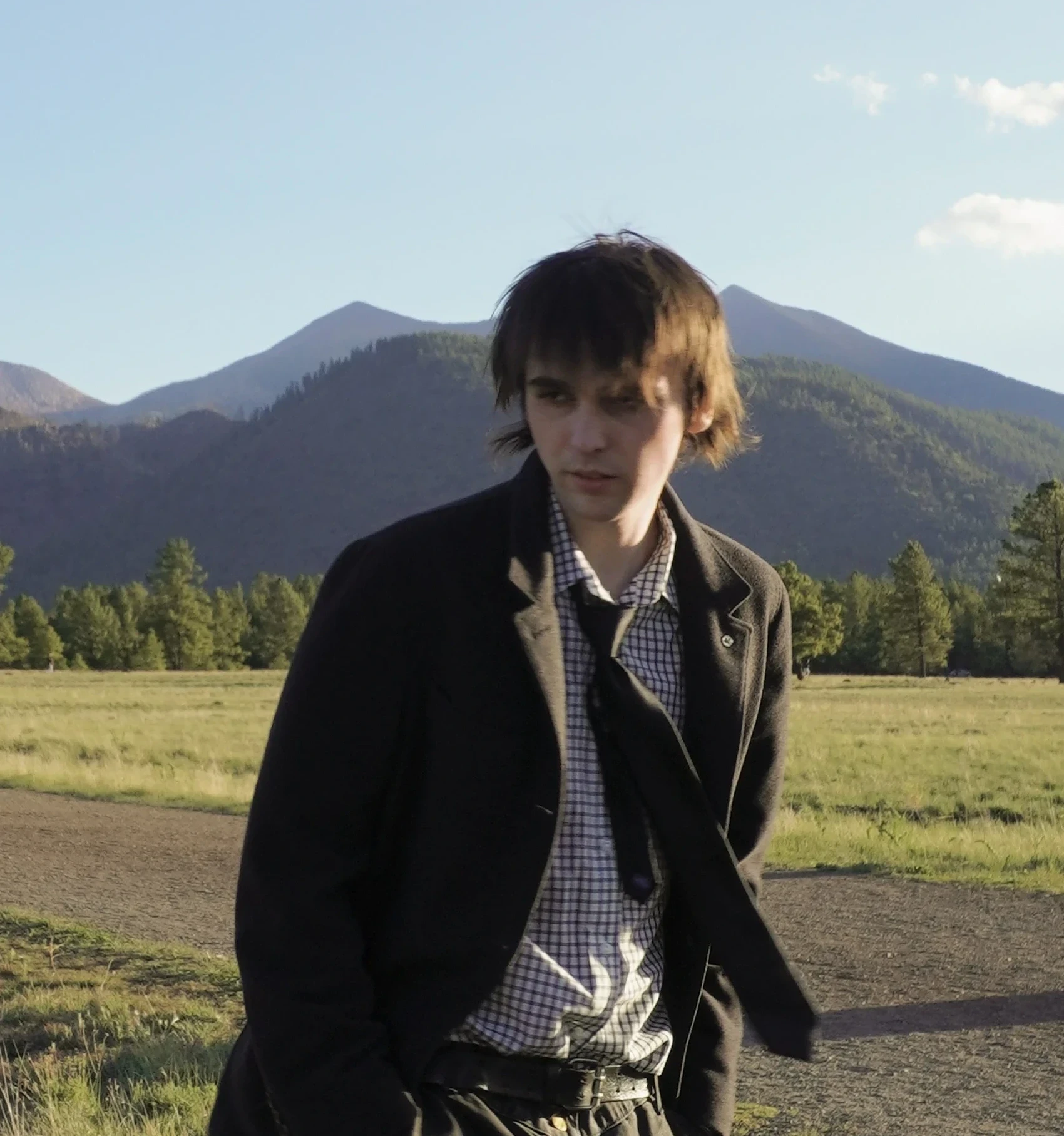 Gavin Schwinn in a dark coat and checked shirt, standing on a dirt path in a grassy meadow with pine trees and the San Francisco Peaks behind him