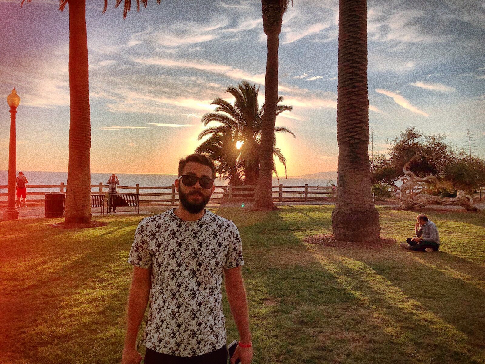 Man in sunglasses and a patterned floral shirt standing between palm trees at sunset in a coastal park, warm sunlight flaring behind him.