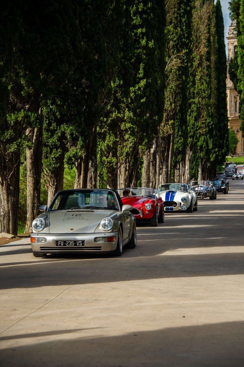 Porsche 911 classique grise en tête d’une file de voitures sous une allée de cyprès, road trip Toscane