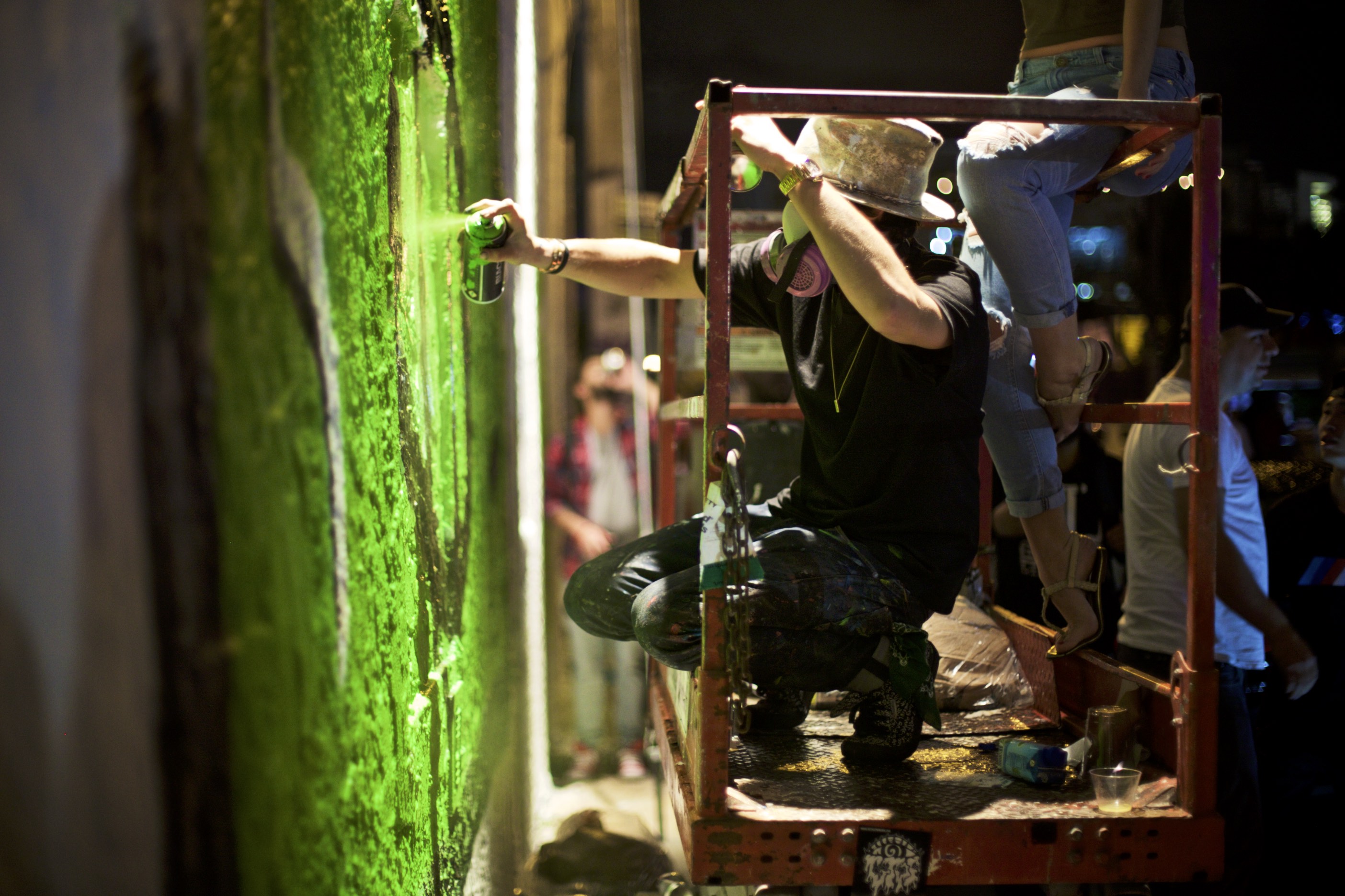 Alec Monopoly painting a large green wall mural from a lift during a live art moment at Art Basel.
