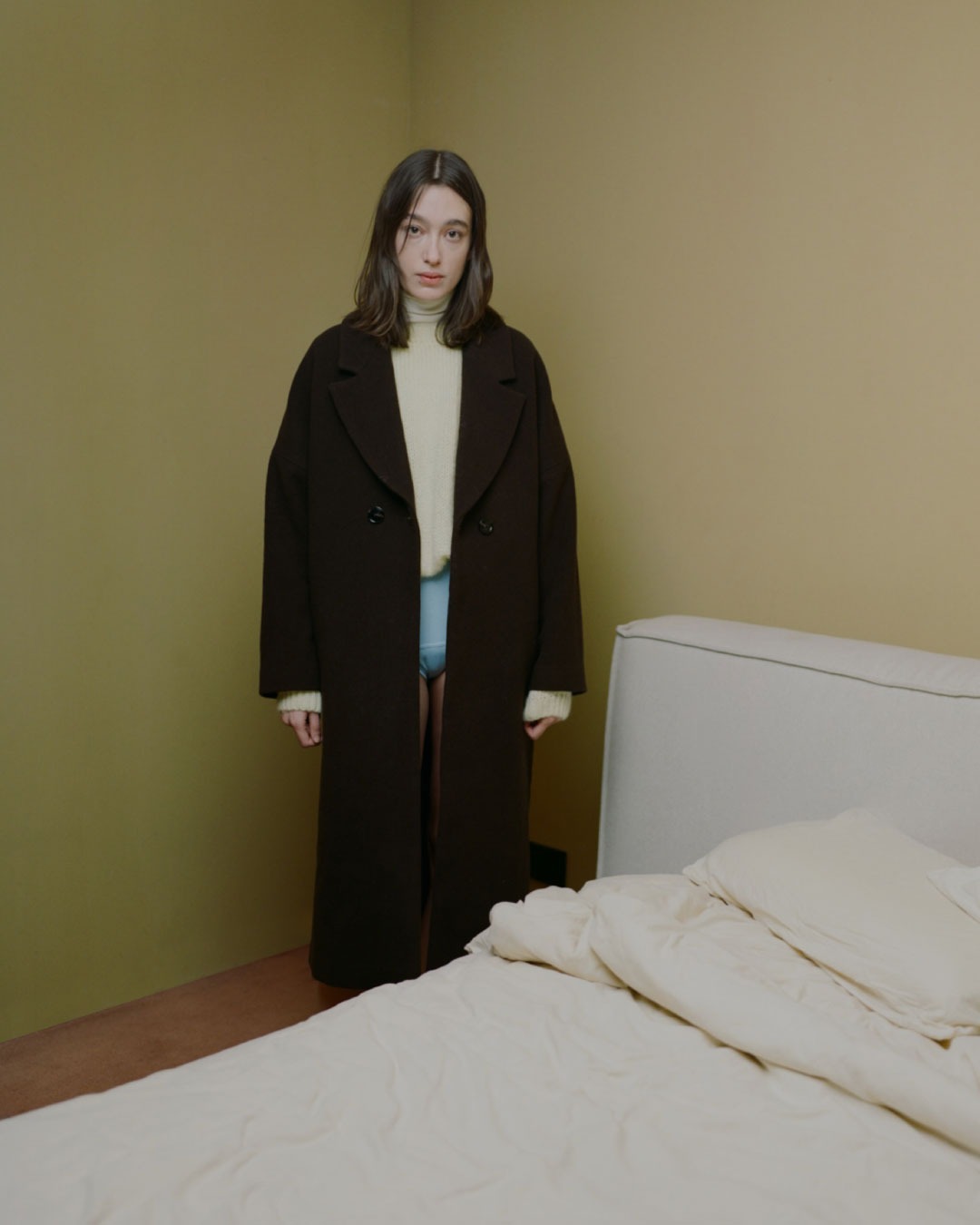 La mannequin Rita Row porte un manteau noiret un haut blanc dans une chambre d'hotel