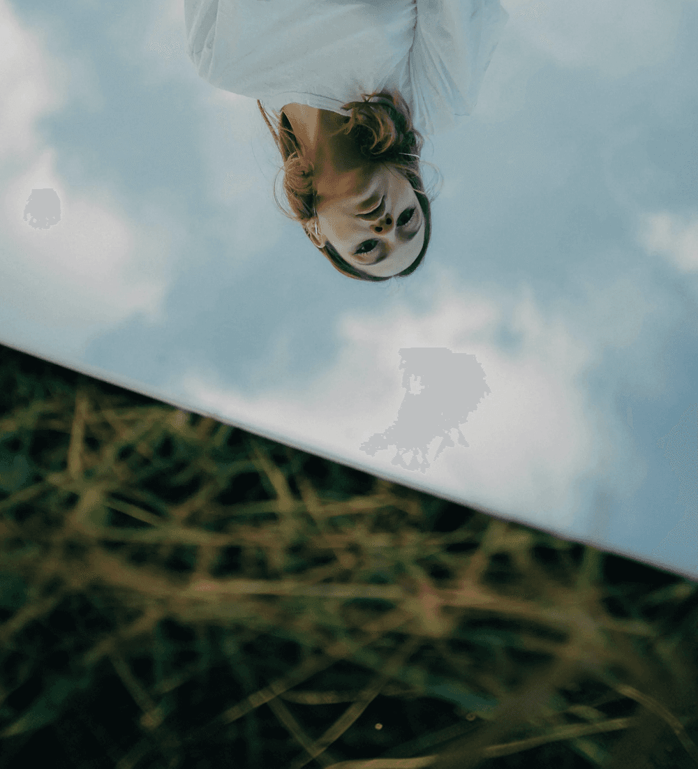An artistic, upside-down reflection in a mirror placed on the ground, showing a person with reddish hair against a cloudy blue sky, with blurry green grass in the foreground.