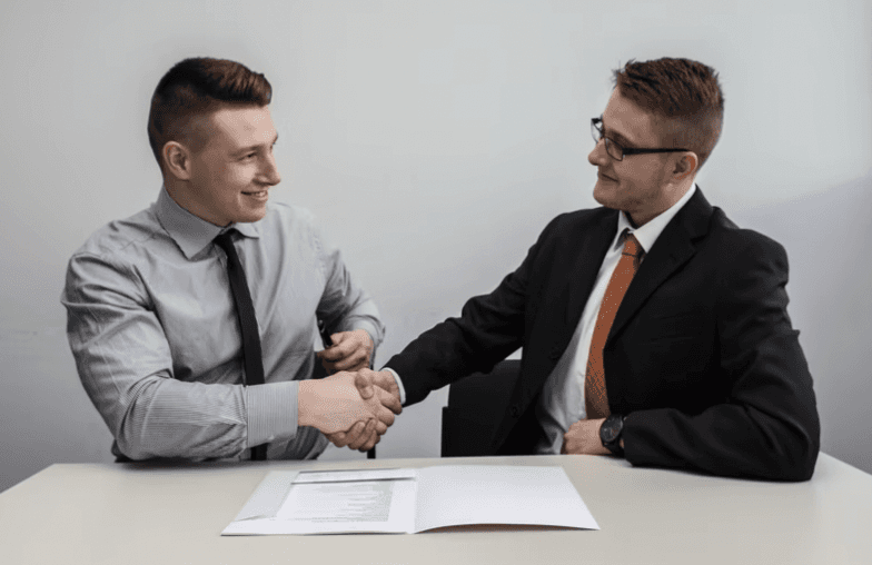 Two men in business attire shaking hands across a desk, with documents in front of them.