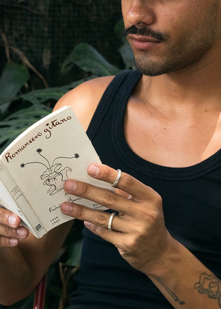 Man holding a book titled 'Romance gátano' with a tattooed arm, surrounded by greenery wearing silver rings.