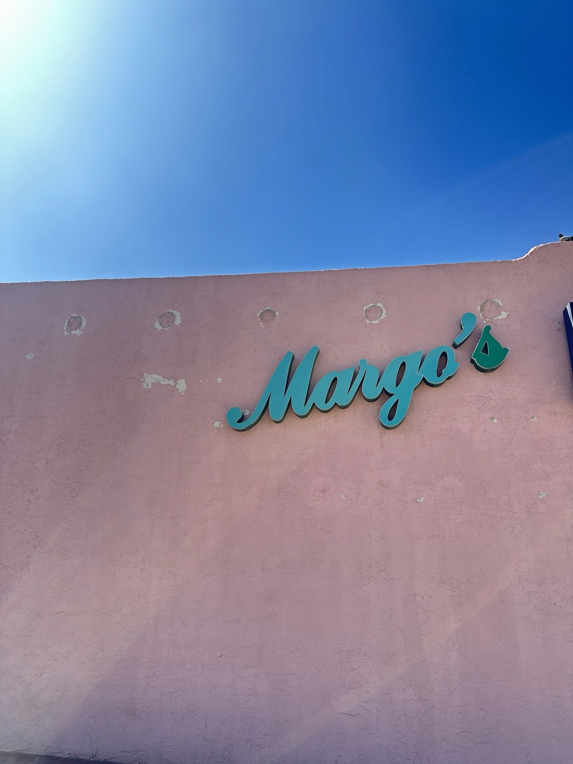 Turquoise Margo’s script lettering on a weathered pink wall under a blue sky