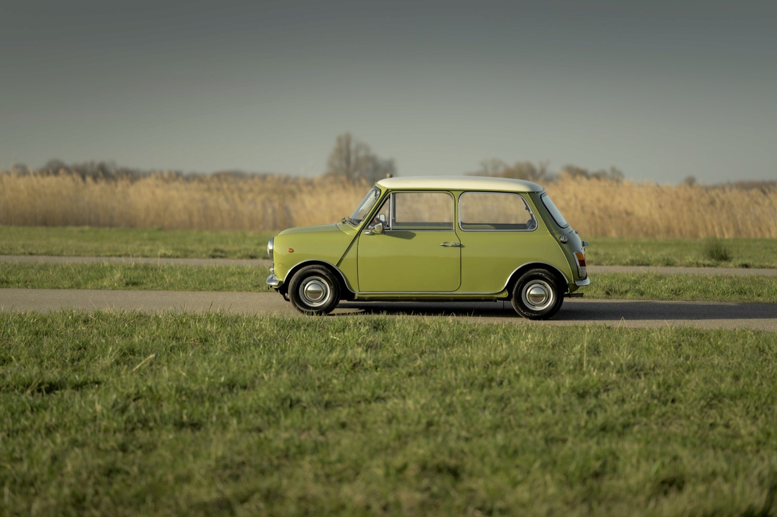 A vintage green mini classic automatic car drives along a road, surrounded by fields and a clear blue sky in the background.