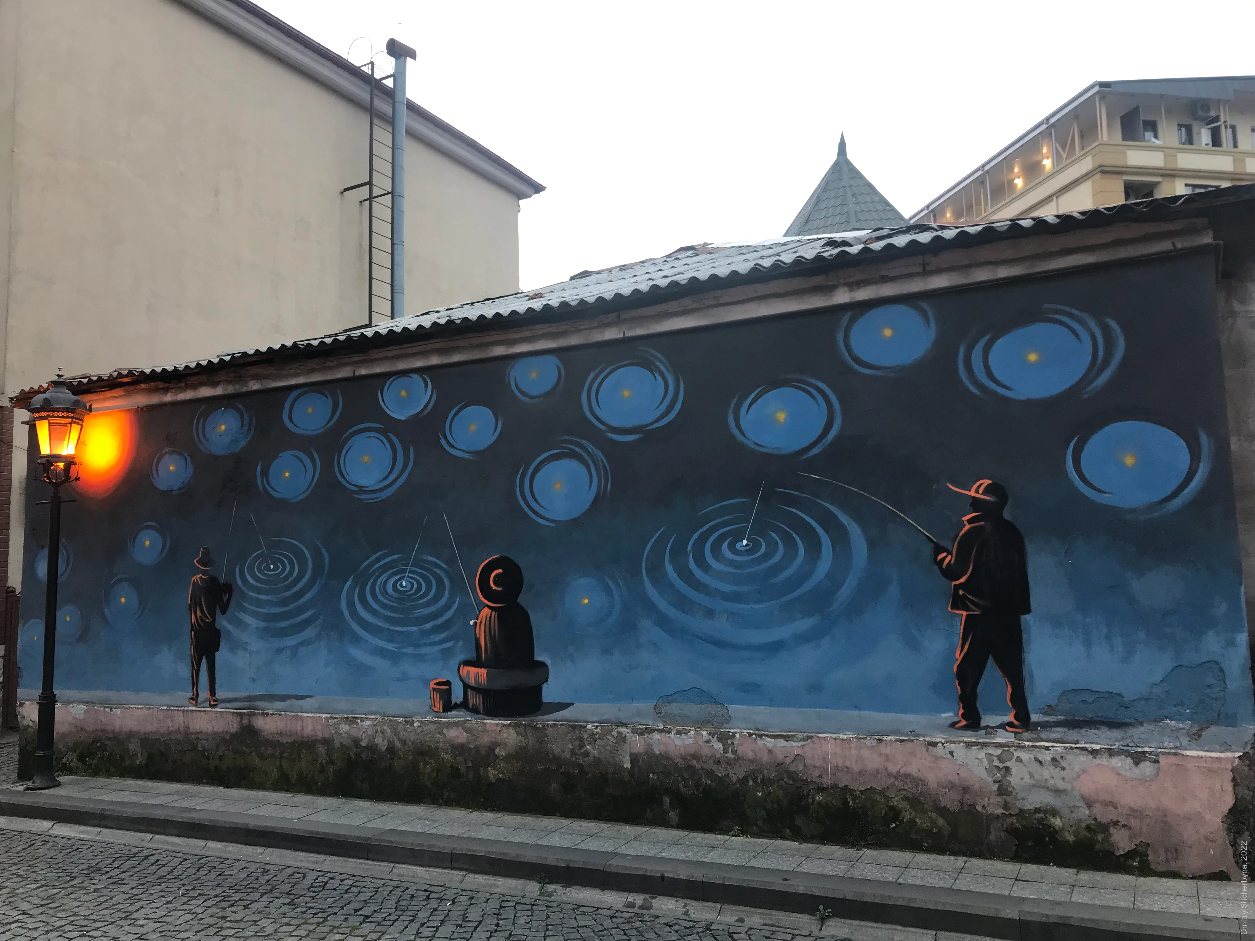A nighttime street mural shows three silhouettes in warm orange tones, fishing with rods in concentric blue ripples that resemble both water and the starry sky. The painting is on a low wall beside a cobblestone street, next to a glowing lamppost.