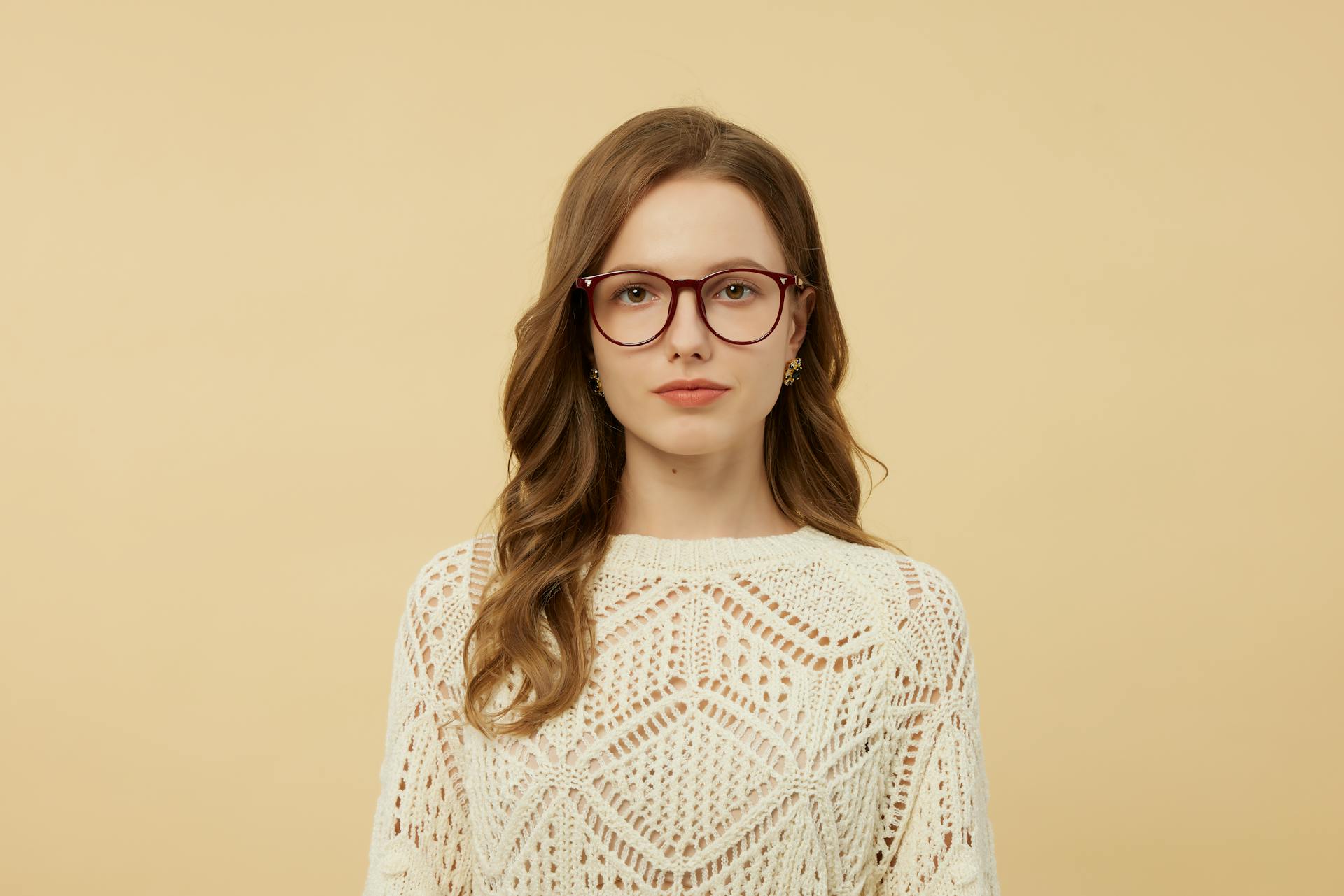 A woman with long brown hair and glasses stands against a pale yellow background, wearing a cream patterned blouse.