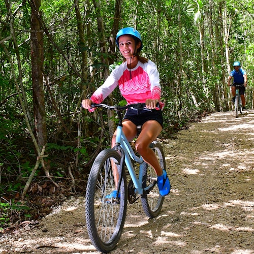 两名戴着头盔的人正在茂密绿色森林中的土路上骑行。前面的人正在微笑。