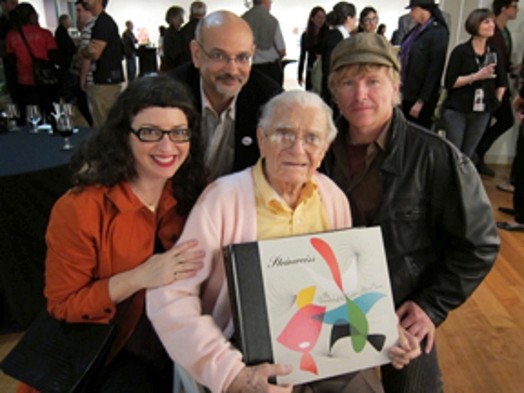 A photo of an old man holding a design book while surrounded by other people.