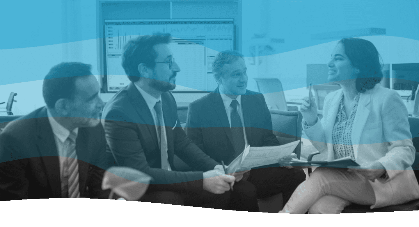 A group of four business professionals engaged in a discussion in an office setting, with charts displayed on the computer screens in the background, suggesting a focus on teamwork and business strategy.