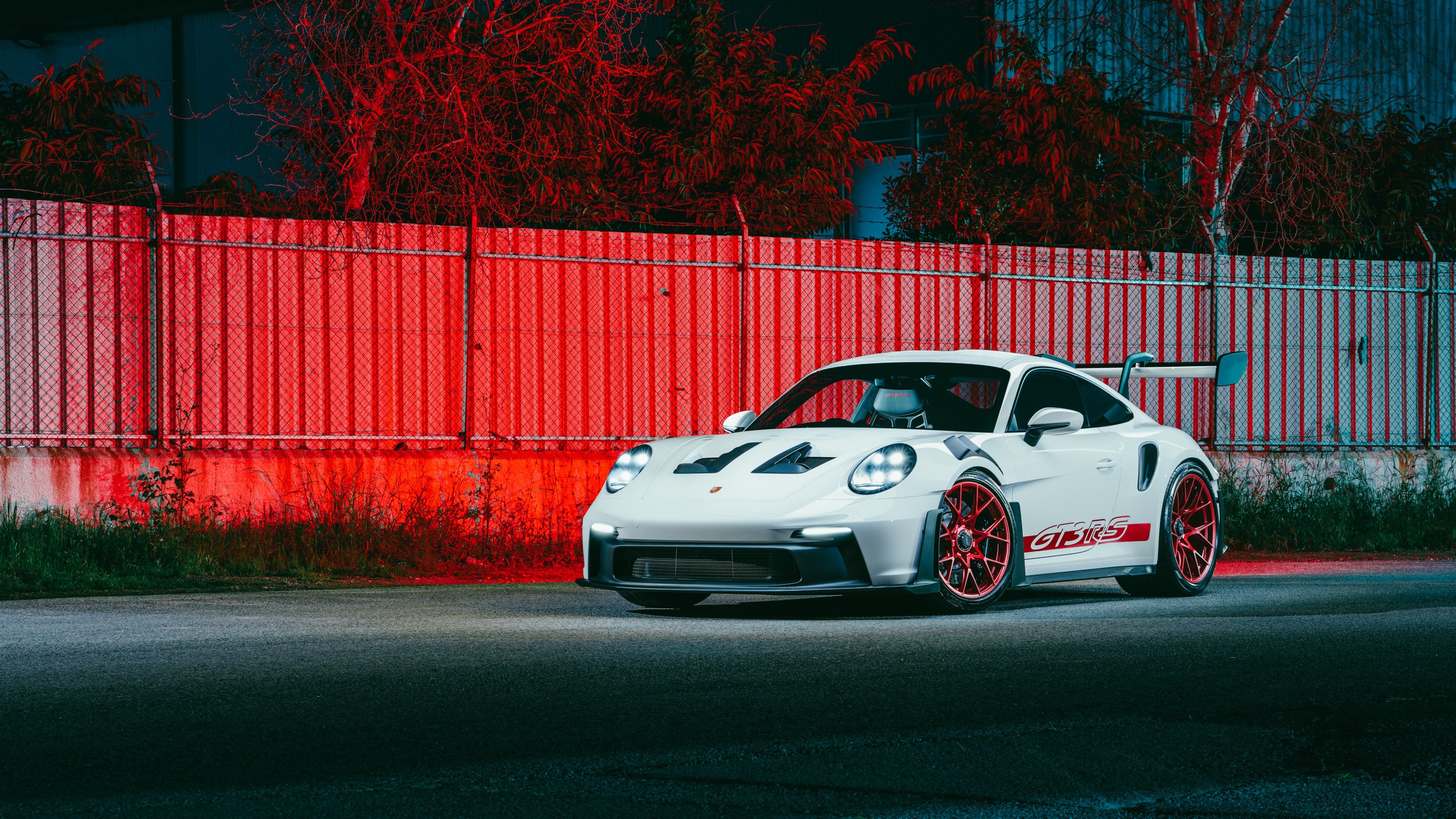 White Porsche 911 GT3 RS (992) with red wheels, photographed from the front three-quarter at night, headlights on, against a red-lit chain-link fence and bare trees, GT3 RS side decal and bonnet vents visible, in an industrial setting in Nicosia.