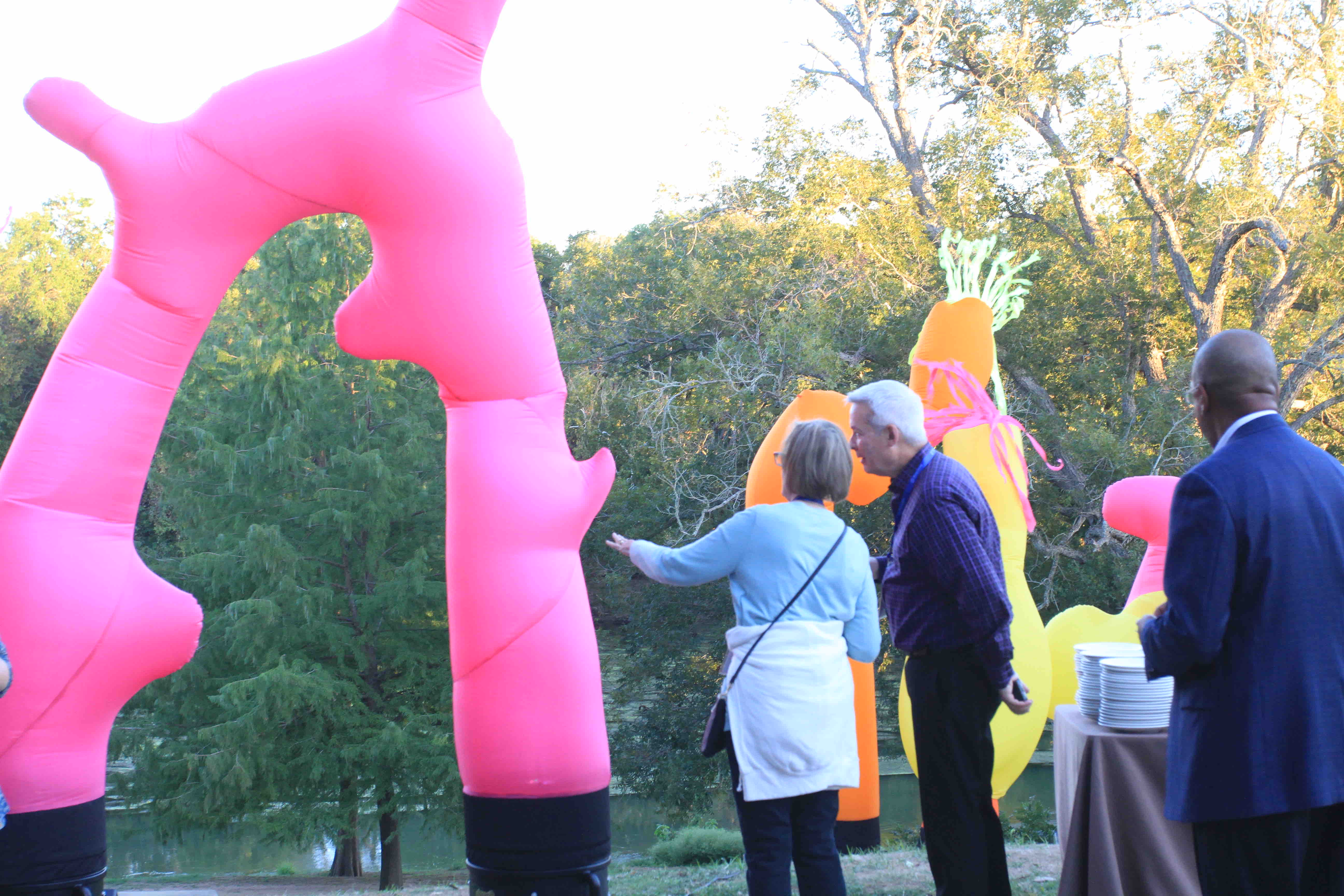 A group of people interact with large pink inflatable sculptures in a park setting. Trees are visible in the background.