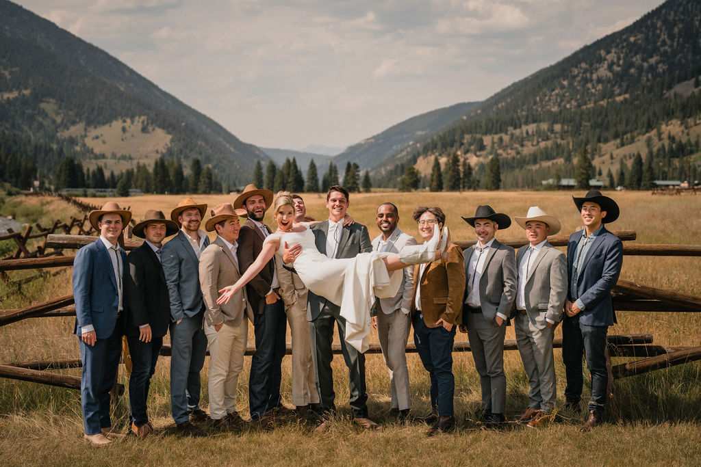 A fun group photo of the wedding party where the groomsmen hold the bride horizontally in the air. Everyone is smiling in front of a rustic wooden fence and mountain landscape.