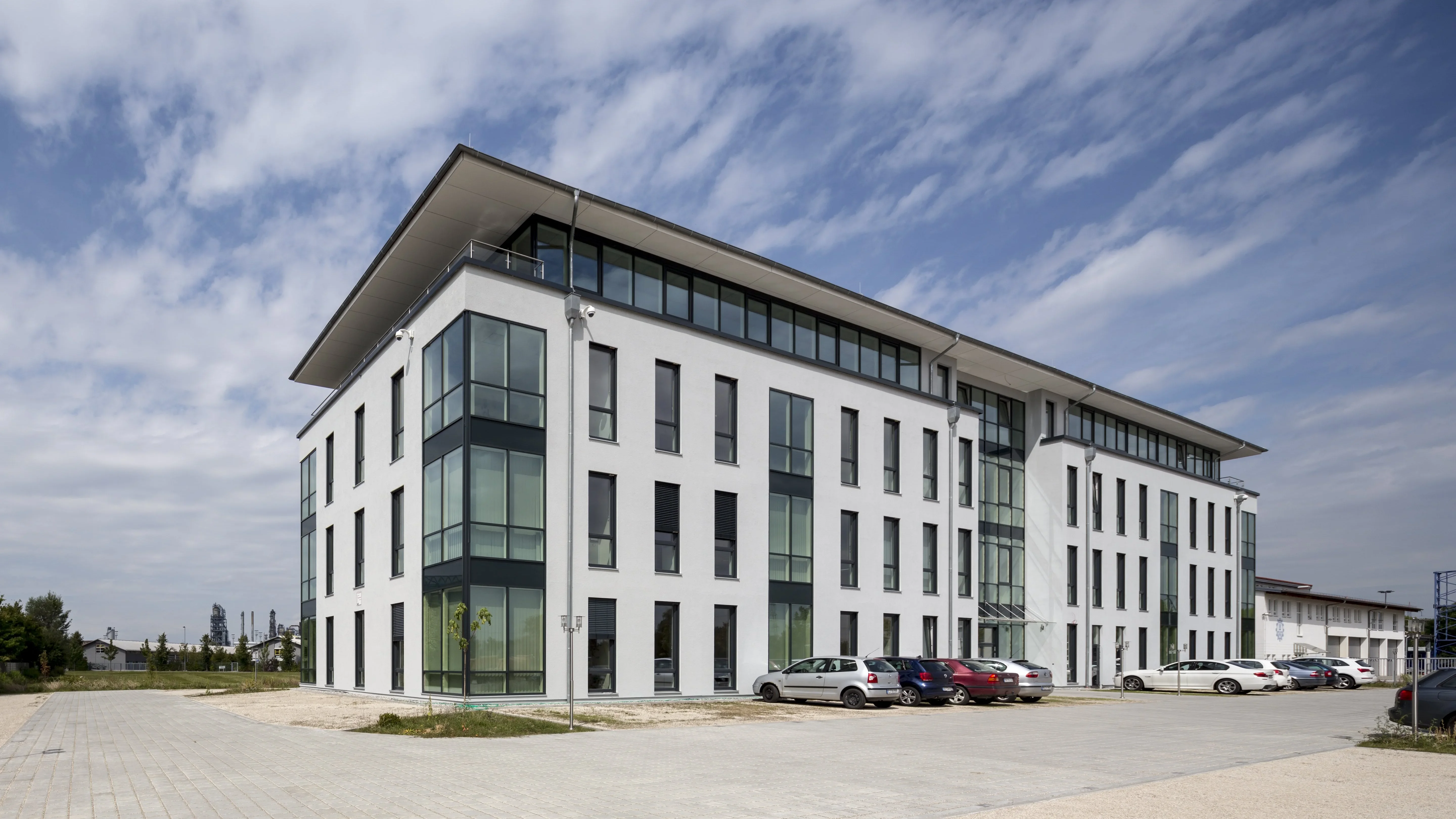 Modernes Bürogebäude mit weißer Fassade und Glasfronten, umgeben von einem Parkplatz mit gepflasterter Fläche und mehreren Autos.