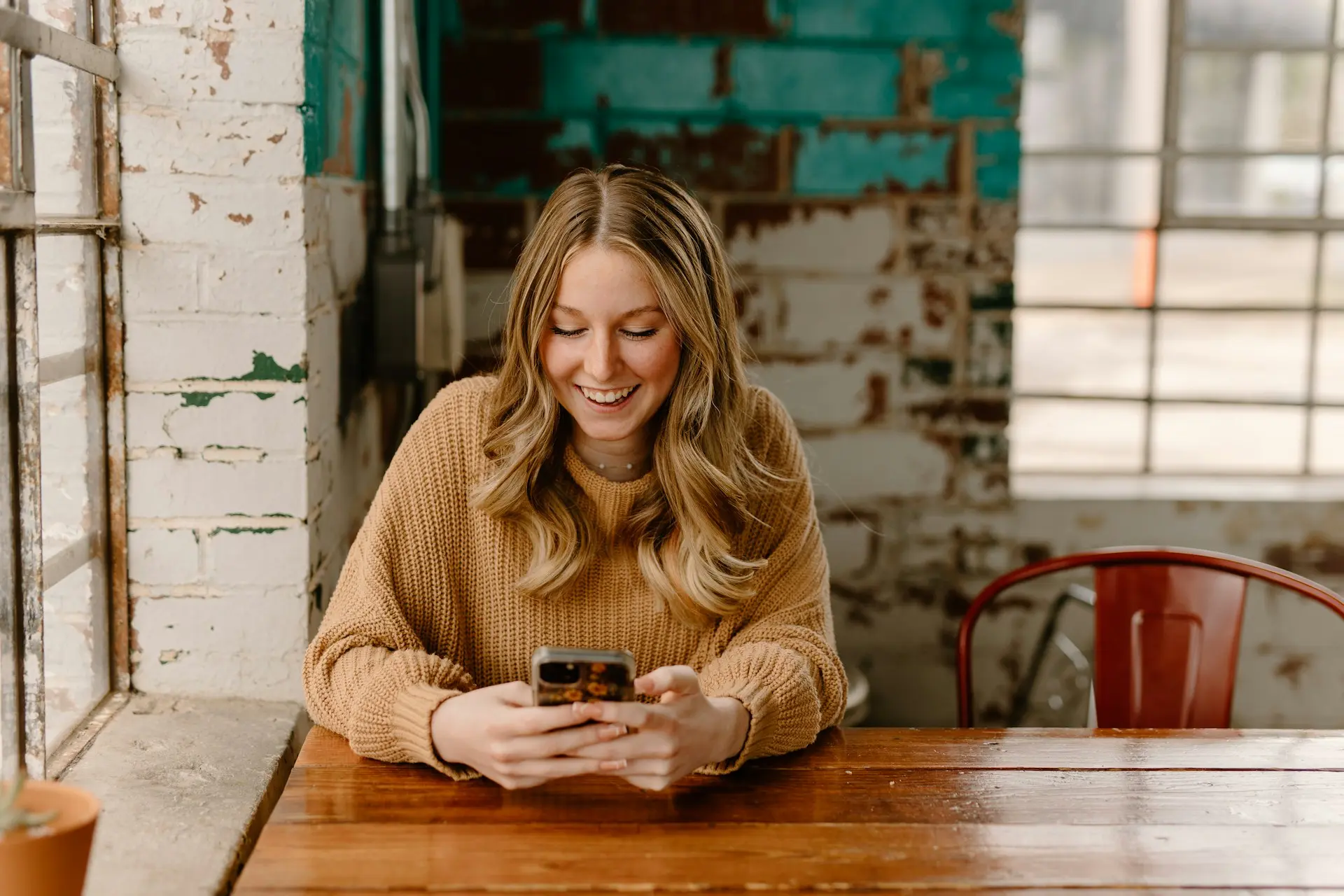 Woman sitting at a wooden table in a café, smiling while using her smartphone near a window with natural light.