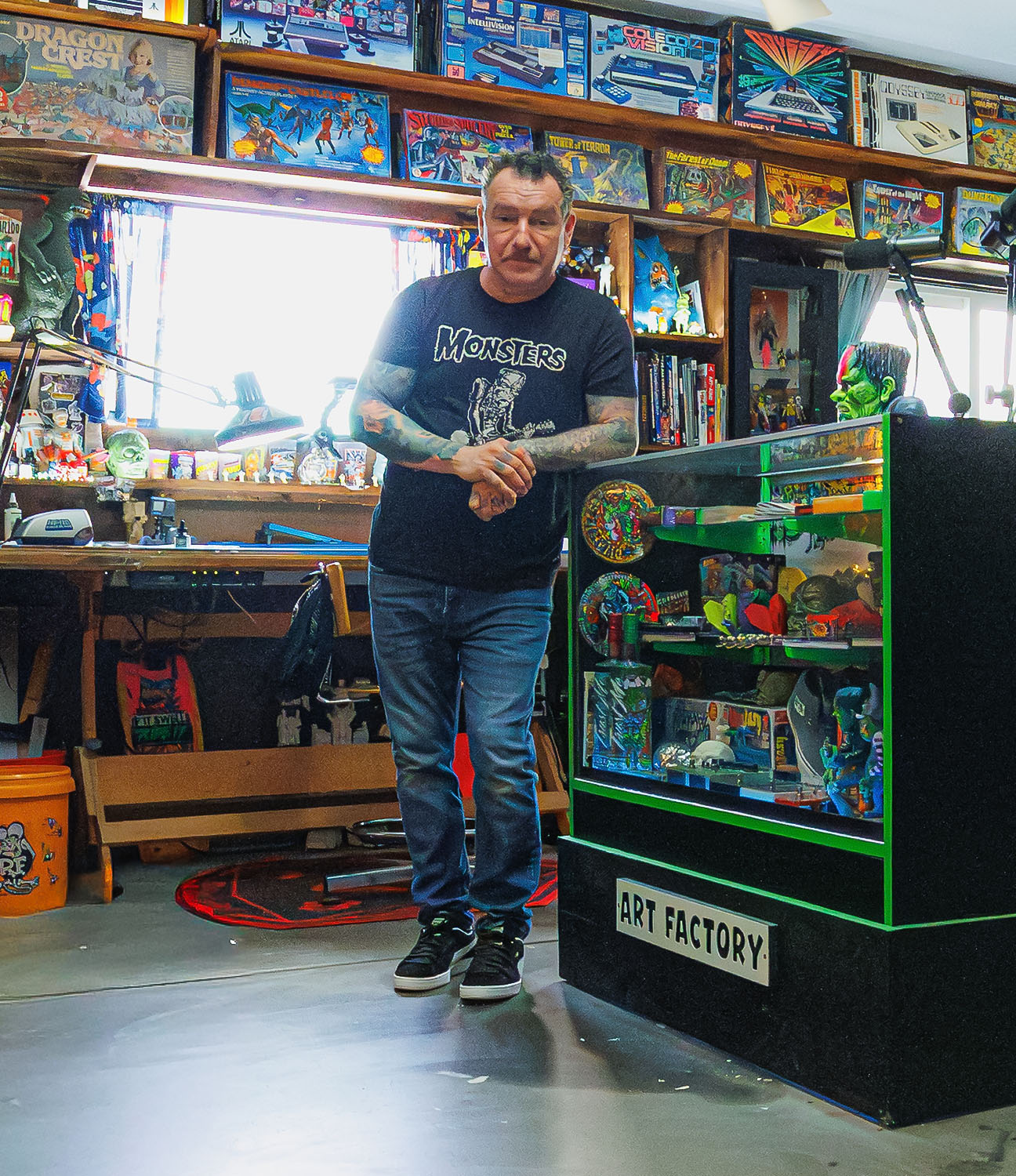 Artist Dirty Donny Gillies standing in his brightly lit studio filled with vintage toys, art supplies, and colorful collectibles, surrounded by shelves packed with models, posters, and pinball machines.