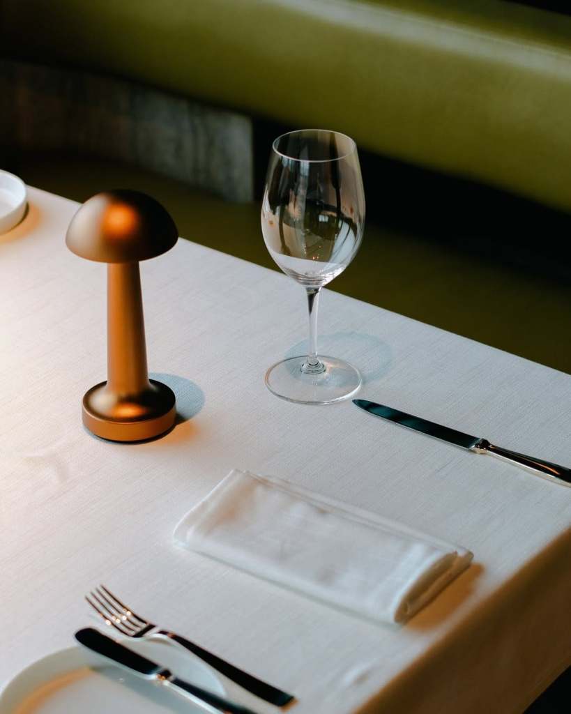 A elegantly set dining table featuring a brown lamp, a wine glass, a white napkin, and silverware on a white tablecloth.