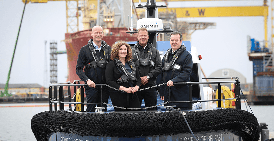 Group of four people wearing safety gear standing on a workboat at an industrial port.