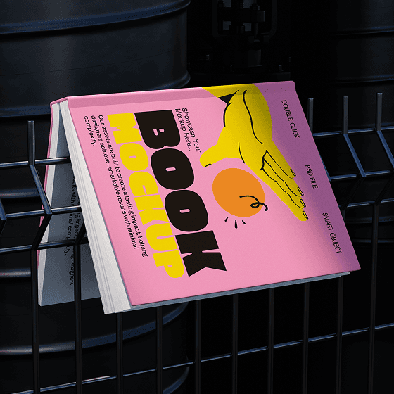 Pink book mockup with bold graphics caught in a dark metal fence in front of black barrels.