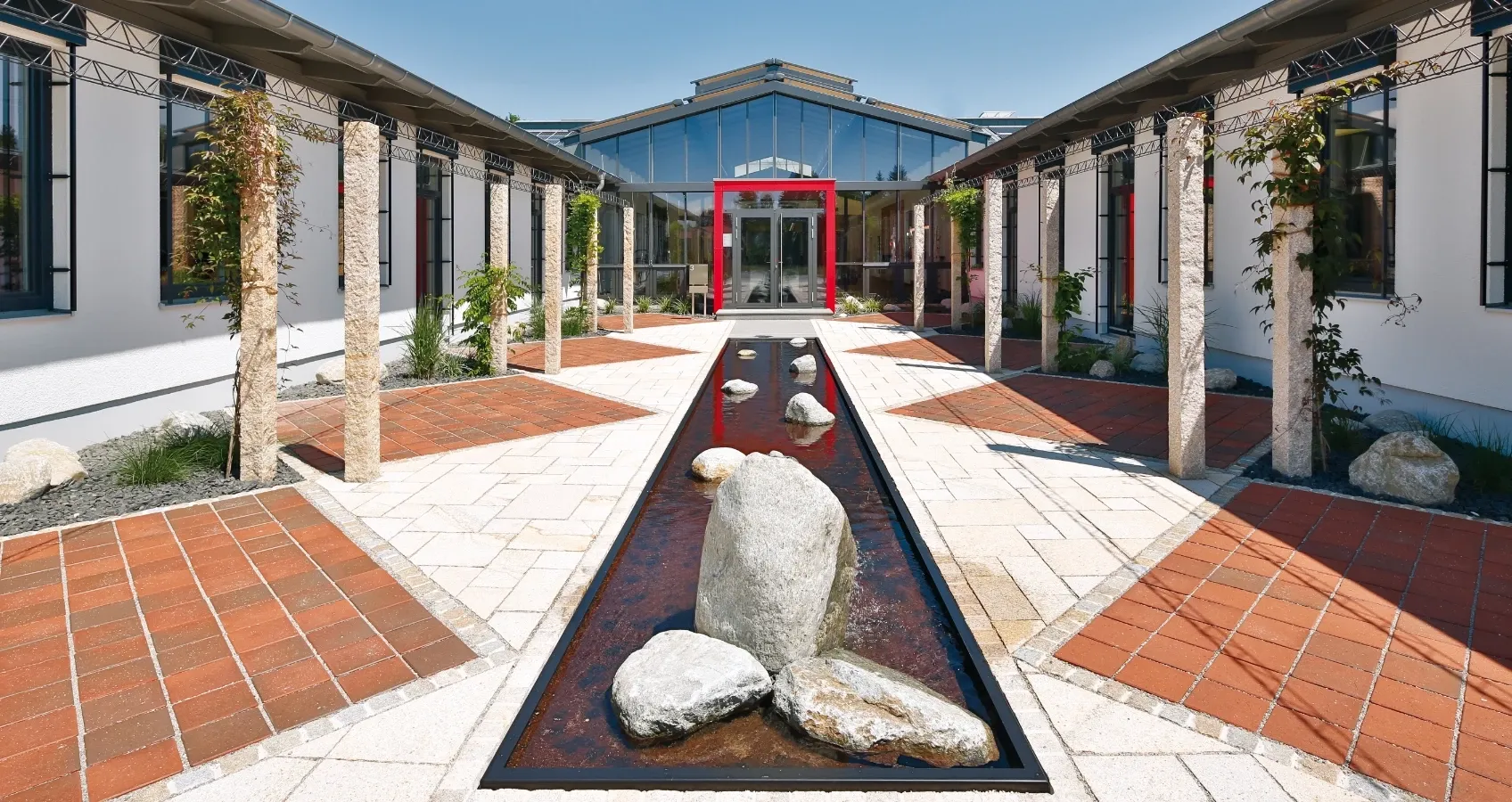 Moderner Innenhof mit Wasserspiel und Steinfiguren, flankiert von Bürogebäuden mit Glasfront.