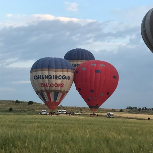 Tatlong hot air balloon sa lupa, isa ay pula at dalawa ay asul, may mga tao at sasakyan malapit sa kanila sa isang madamong parang sa ilalim ng maulap na kalangitan.