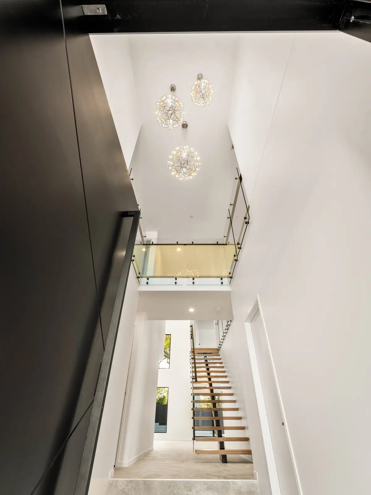 A modern multi-story interior features a wooden staircase, glass railings, and spherical chandeliers.