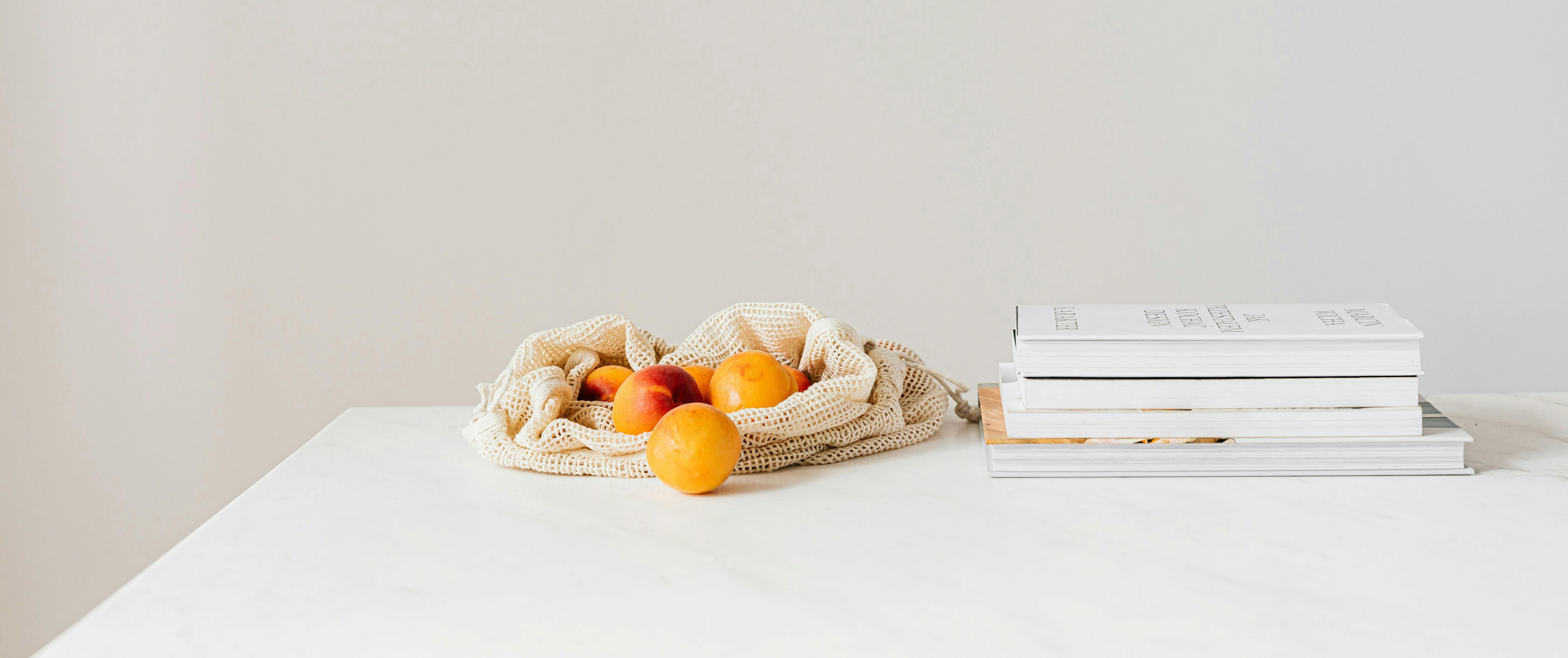 A bag of oranges and a stack of white books on a white table