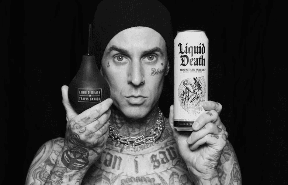 Una persona con una camiseta negra y pantalones de color claro está de pie en la cima rocosa de una montaña al atardecer, sosteniendo una cámara frente a su cara para tomar una foto. Un mar de nubes se extiende detrás de ellos, iluminado por la luz dorada del sol poniente.