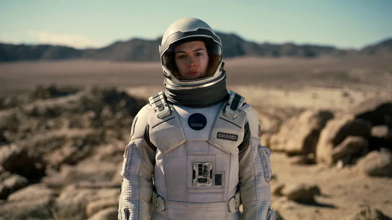 An astronaut in a white spacesuit with a visible NASA logo stands in a barren, rocky desert landscape under a clear blue sky.