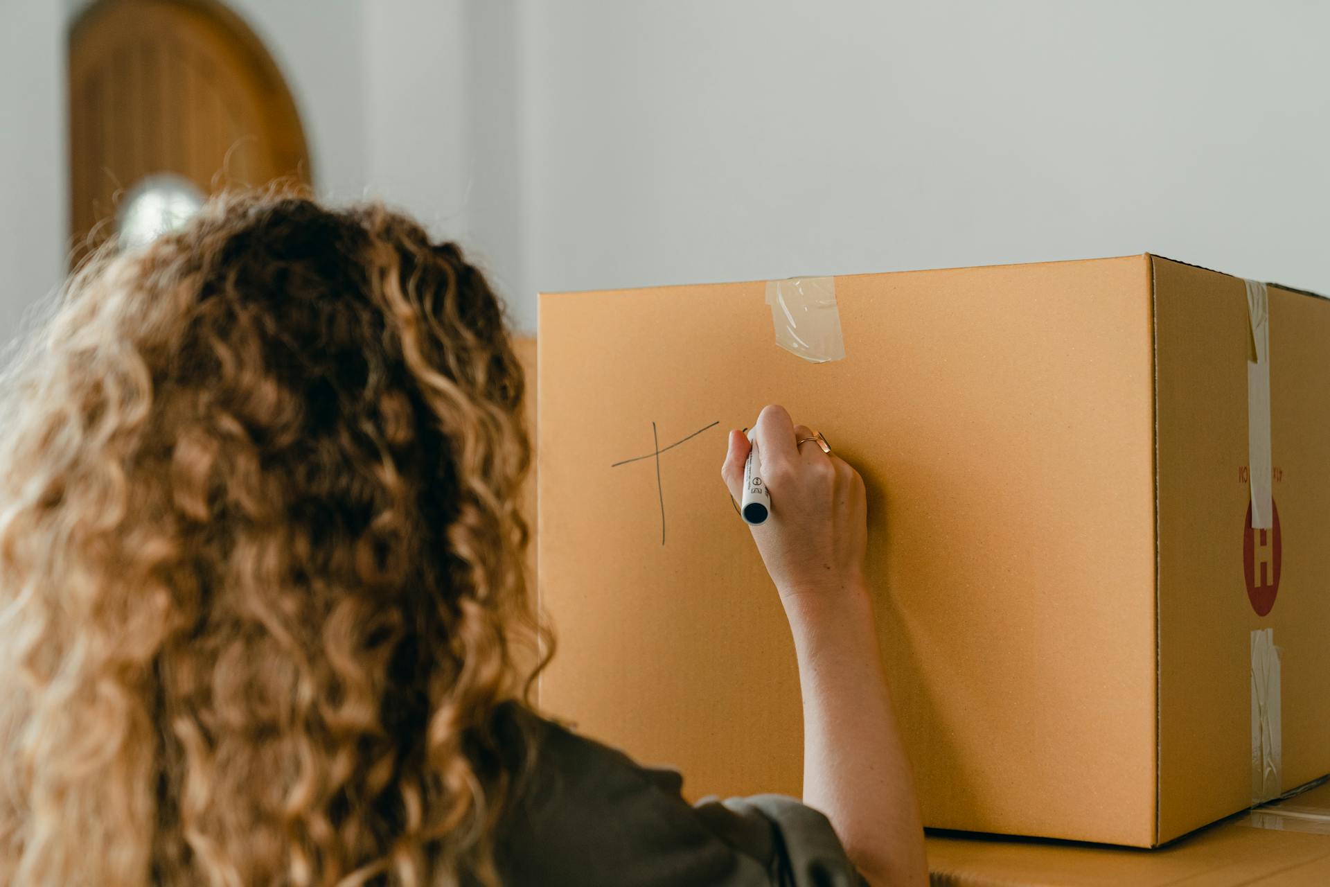 A person with long, curly, brown hair is seen from behind, writing on a plain brown cardboard moving box with a black marker.