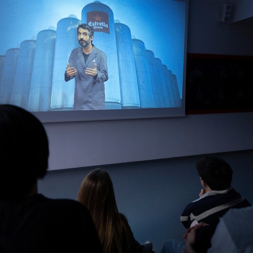 几个人正在观看一位男士在投影屏幕上讲话，背景中有大型筒仓。