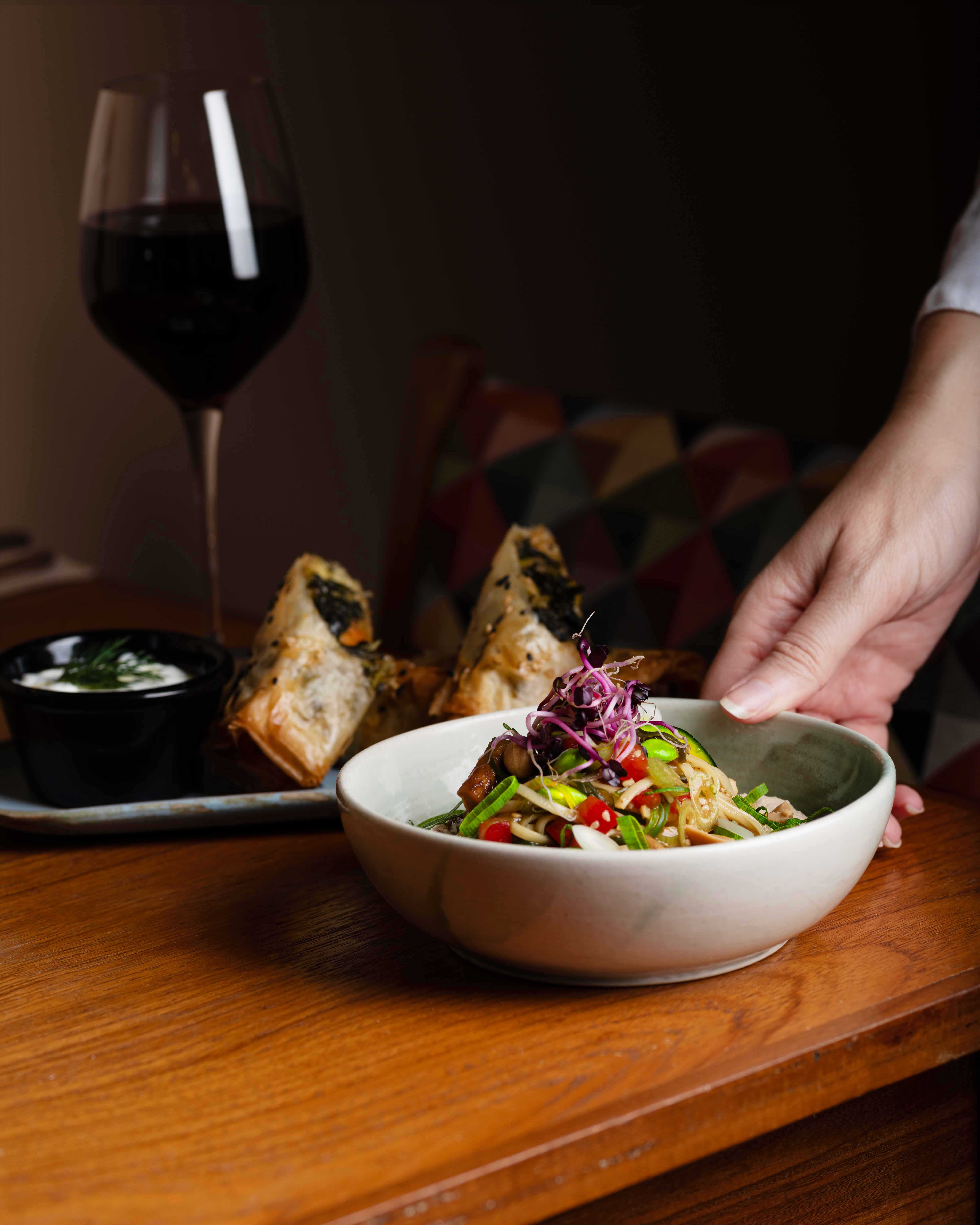 Bol de fideos vegetarianos con rollitos y vino tinto natural en Faire. Tapas & Wine