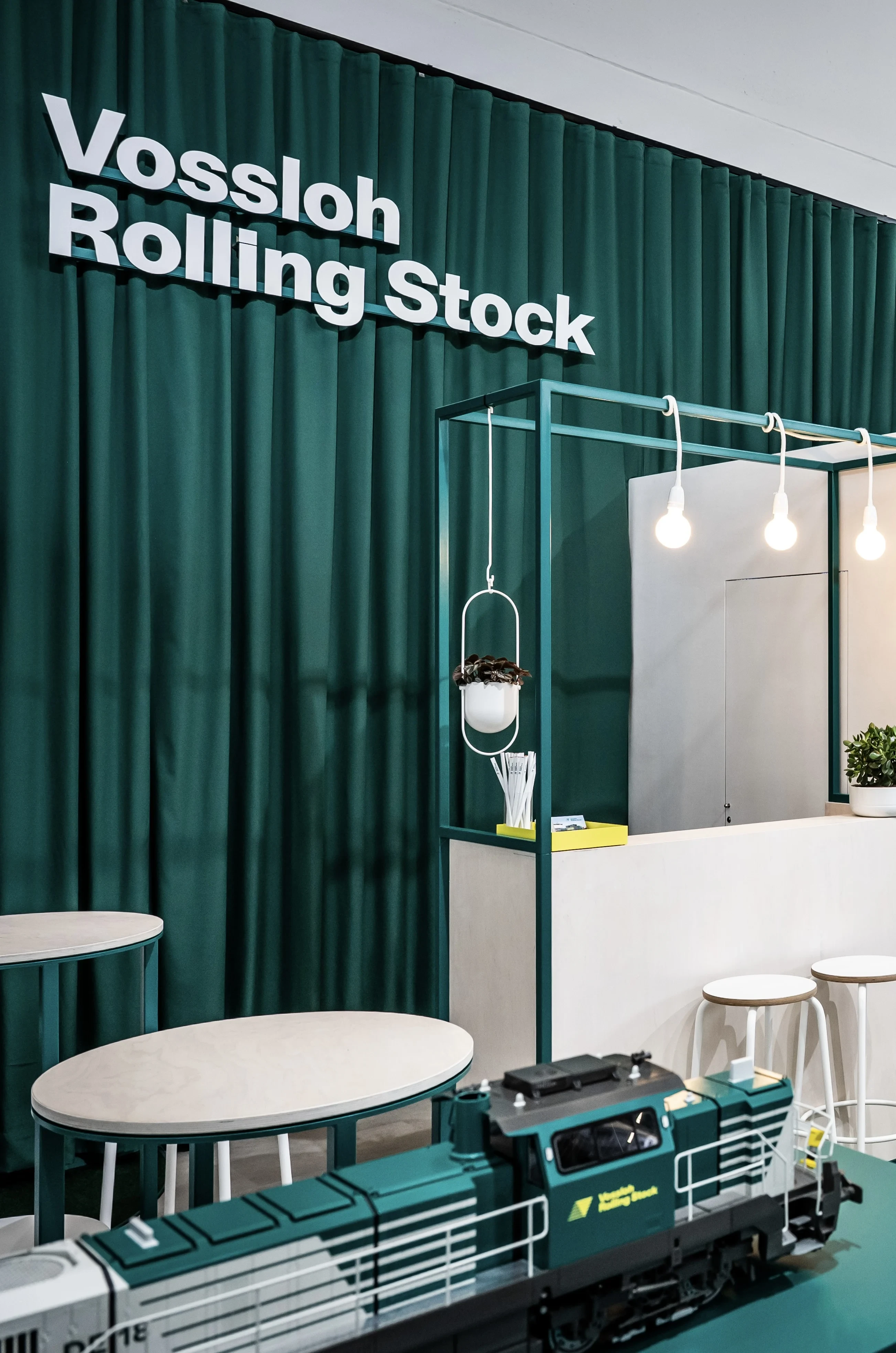 Vossloh Rolling Stock lettering on green curtain above a model locomotive and white counter with pendant lights.
