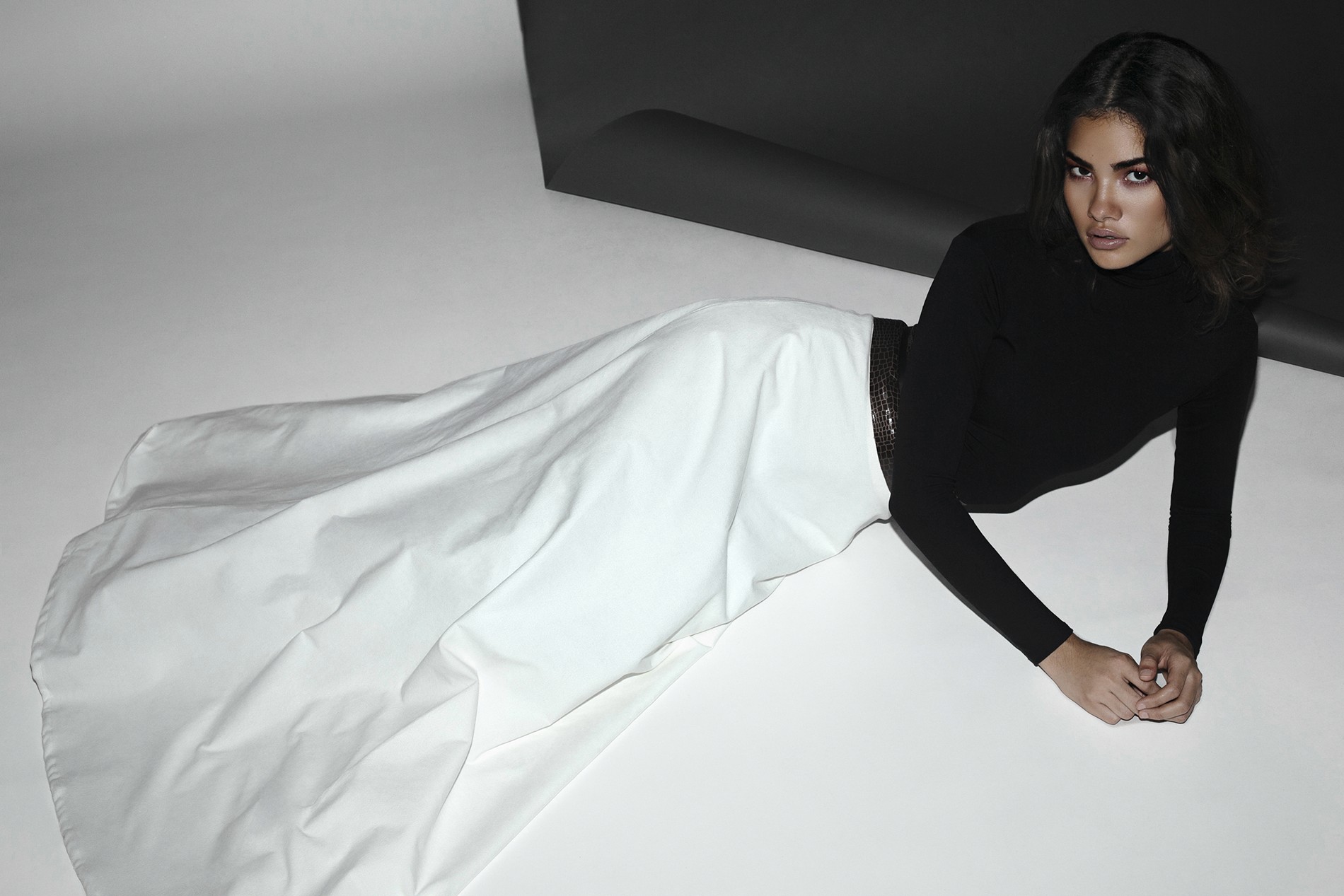 Cenit lying on a white floor in a black turtleneck and white skirt by Jean Cintron, creating a striking visual contrast.